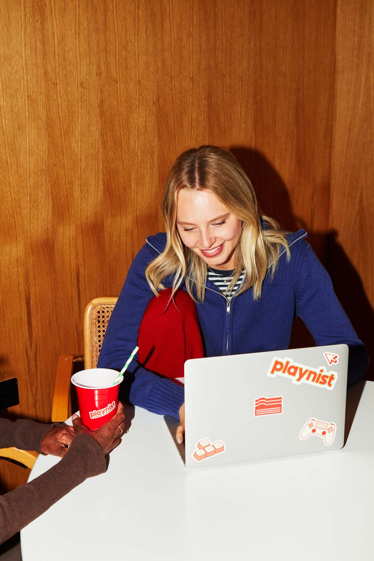 Smiling blonde woman in blue cardigan using laptop with Playnist gaming stickers while holding red branded cup, sitting at wooden table against wood-paneled wall.